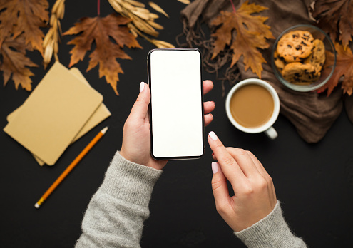 Technology and season concept. Woman holding cellphone with blank screen over autumn fallen leaves background