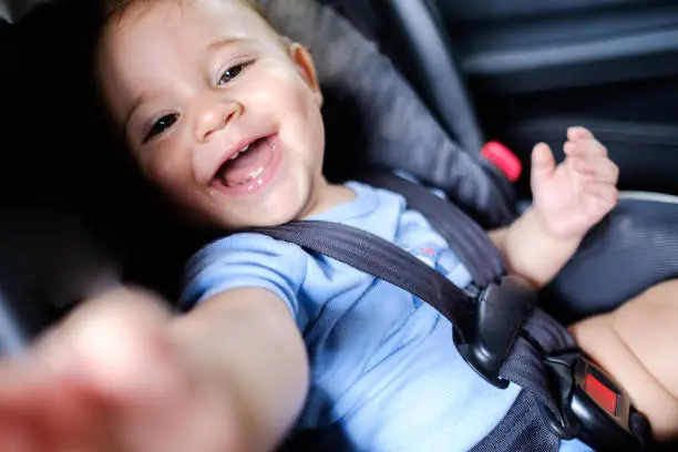 Baby boy in baby comfort in car, looking at camera and smiling showing his teeth and trying to catch the camera