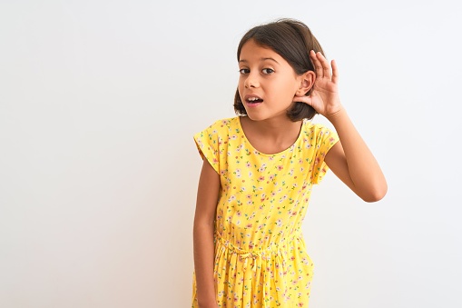 Young beautiful child girl wearing yellow floral dress standing over isolated white background smiling with hand over ear listening an hearing to rumor or gossip. Deafness concept.