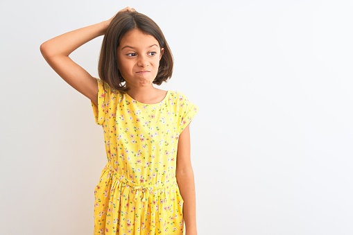 Young beautiful child girl wearing yellow floral dress standing over isolated white background confuse and wondering about question. Uncertain with doubt, thinking with hand on head. Pensive concept.