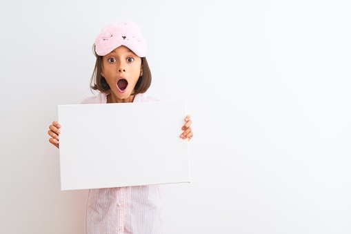 Child girl wearing sleep mask and pajama holding banner over isolated white background scared in shock with a surprise face, afraid and excited with fear expression