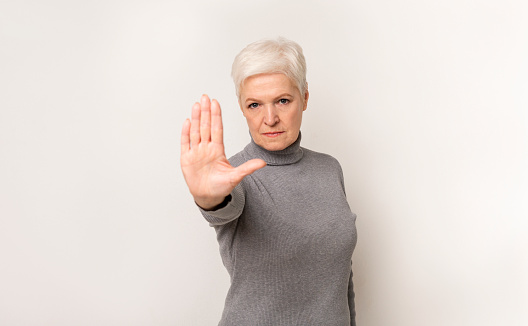 No means no. Portrait of serious elderly woman showing stop gesture standing with stretched hand, light studio background with copy space