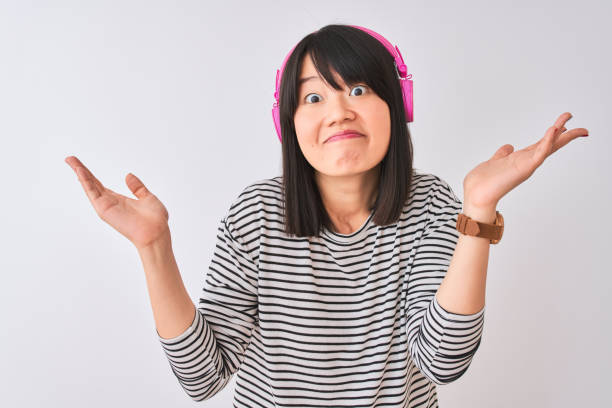 chinese woman listening to music using pink headphones over isolated white background clueless and confused expression with arms and hands raised. doubt concept. - head and shoulders audio imagens e fotografias de stock