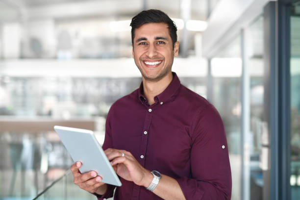 I do all my work on this device Cropped portrait of a handsome young businessman using a digital tablet while standing in modern office indian young man stock pictures, royalty-free photos & images