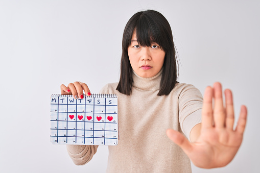 Young beautiful Chinese woman holding menstruation calendar over isolated white background with open hand doing stop sign with serious and confident expression, defense gesture