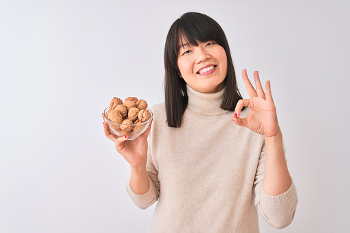 Young beautiful Chinese woman holding bowl with walnuts over isolated white background doing ok sign with fingers, excellent symbol