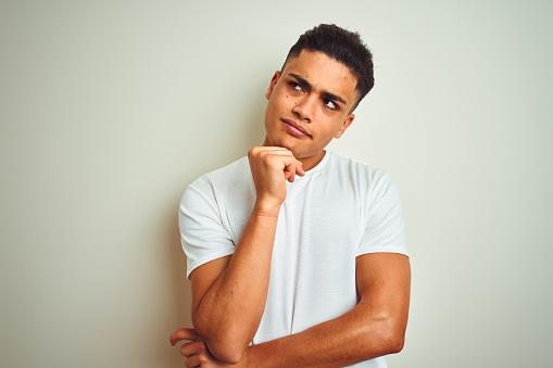 Young brazilian man wearing t-shirt standing over isolated white background with hand on chin thinking about question, pensive expression. Smiling with thoughtful face. Doubt concept.
