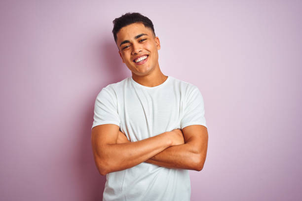 고립된 분홍색 배경 위에 서있는 티셔츠를 입은 젊은 브라질 남자가 카메라를 바라보며 두 팔을 교차하고 미소짓고 있는 행복한 얼굴. 긍정적 인 사람. - 청년 남자 뉴스 사진 이미지