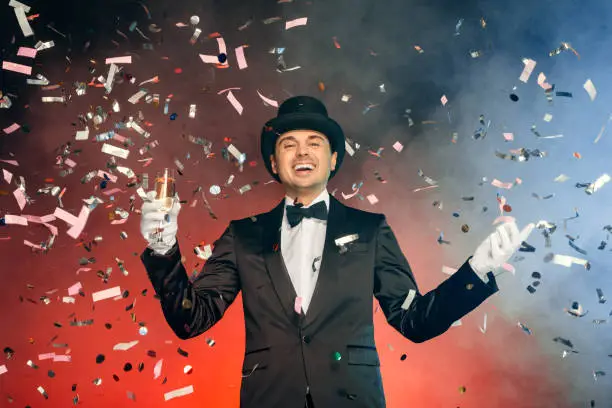 Professional showman wearing suit top hat and gloves standing isolated on blue and red background flying confetti holding glass of champagne laughing happy
