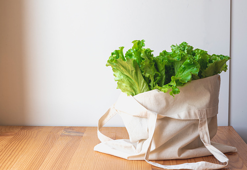 Sustainable lifestyle concept. Cotton eco bag with green lettuce stands on a wooden table, white background. Free plastic, zero waste concept.