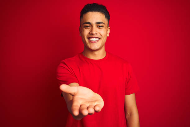 Young brazilian man wearing t-shirt standing over isolated red background smiling cheerful offering palm hand giving assistance and acceptance. Young brazilian man wearing t-shirt standing over isolated red background smiling cheerful offering palm hand giving assistance and acceptance. hand palm stock pictures, royalty-free photos & images