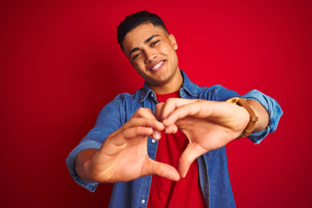 camisa desgastando do denim do homem brasileiro novo que está sobre o fundo vermelho isolado que sorri no amor que mostra o símbolo e a forma do coração com mãos. conceito romântico. - jovem no coração - fotografias e filmes do acervo