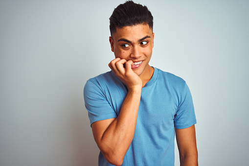 Young brazilian man wearing blue t-shirt standing over isolated white background looking stressed and nervous with hands on mouth biting nails. Anxiety problem.