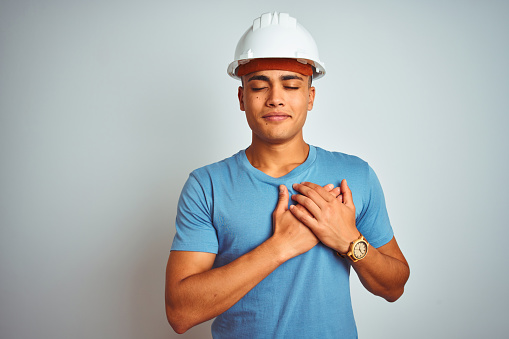 Young brazilian engineer man wearing security helmet standing over isolated white background smiling with hands on chest with closed eyes and grateful gesture on face. Health concept.