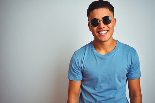 Young brazilian man wearing blue t-shirt and sunglasses over isolated white background with a happy and cool smile on face. Lucky person.