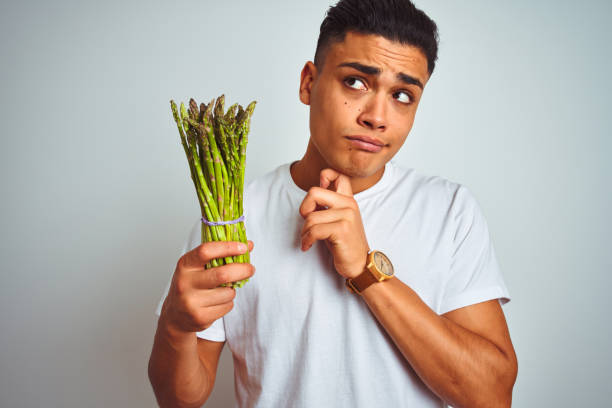 Young brazilian man eating asparagus standing over isolated grey background serious face thinking about question, very confused idea Young brazilian man eating asparagus standing over isolated grey background serious face thinking about question, very confused idea eating asparagus stock pictures, royalty-free photos & images