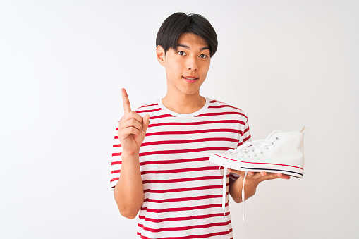 Handsome chinese man holding casual sneakers standing over isolated white background surprised with an idea or question pointing finger with happy face, number one