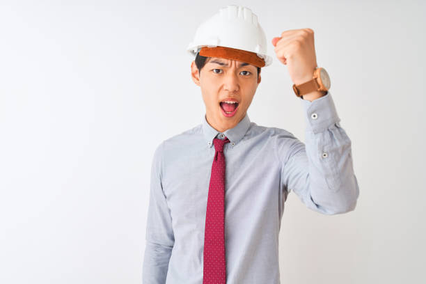 istock Chinese architect man wearing tie and helmet standing over isolated white background angry and mad raising fist frustrated and furious while shouting with anger. Rage and aggressive concept. 1174459551