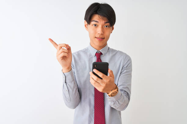 istock Chinese businessman wearing tie using smartphone standing over isolated white background very happy pointing with hand and finger to the side 1174459763