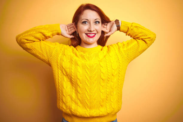 hermosa mujer pelirroja usando suéter de invierno de pie sobre el fondo amarillo aislado sonriendo orejas tirando de los dedos, gesto divertido. problema de audición - bizarre women portrait pierced fotografías e imágenes de stock