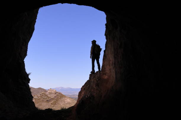 istock EXPLORER WITH HAT AND BACKPACK UP ON A SHELF IN A LARGE CAVE WATCHING A CASTLE IN THE DISTANCE 1174463120