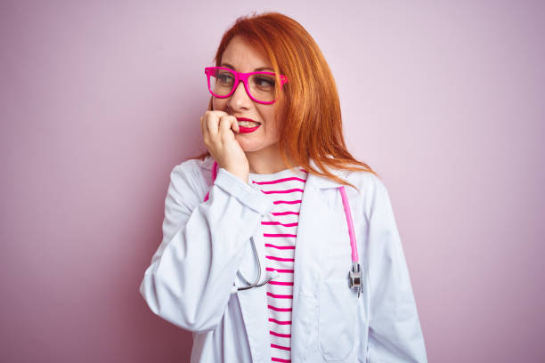 istock Young redhead doctor woman using stethoscope standing over isolated pink background looking stressed and nervous with hands on mouth biting nails. Anxiety problem. 1174466789