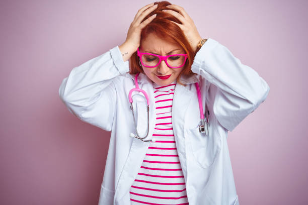 istock Young redhead doctor woman using stethoscope standing over isolated pink background suffering from headache desperate and stressed because pain and migraine. Hands on head. 1174466801