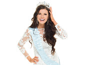 One person / waist up of 20-29 years old adult beautiful brown hair / long hair caucasian female / young women beauty queen / miss universe pageant in front of white background wearing sash / dress who is successful / smiling / happy / cheerful
