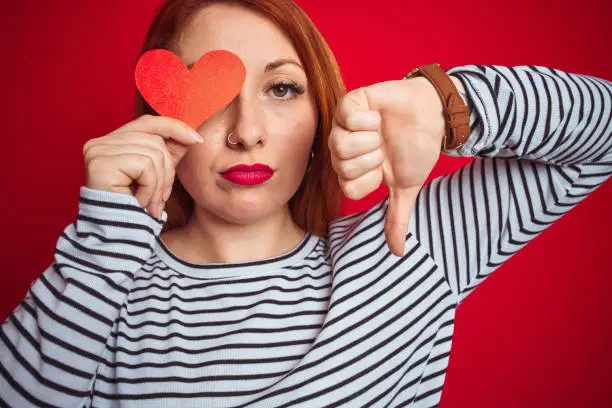 Photo of Young redhead romantic woman holding heart over red isolated background with angry face, negative sign showing dislike with thumbs down, rejection concept