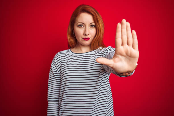 istock Young redhead woman wearing strapes navy shirt standing over red isolated background doing stop sing with palm of the hand. Warning expression with negative and serious gesture on the face. 1174468558