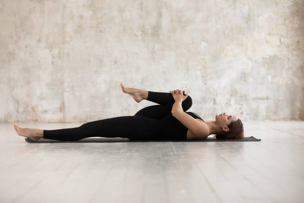 Woman lying on mat doing Half Knees to Chest Pose Woman wear black sport clothes lying on floor practising asana do Half Knees to Chest Pose near grunge wall beige textured background, help ease back pain, flexible body stretch for beginners concept chest torso photos stock pictures, royalty-free photos & images