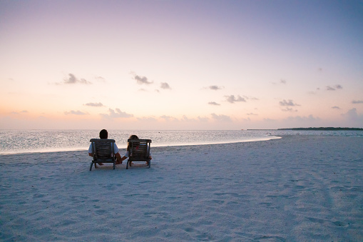Couple enjoying holidays sitting on lounge chairs at tropical paradise beach at Dhiffushi Holiday island at South Ari atoll, Maldives. Beautiful turquoise Indian ocean sea with white sand. Luxury travel holidays background. Model and Property released.