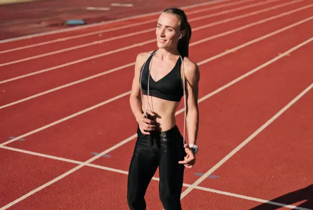 Young fit woman in sportswearwith jumping rope posing on track of stadium with red coating at sunny bright day