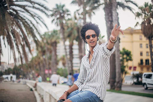 Handsome, young man with afro hairstyle sitting outdoors, listening to music on smart phone and waving.