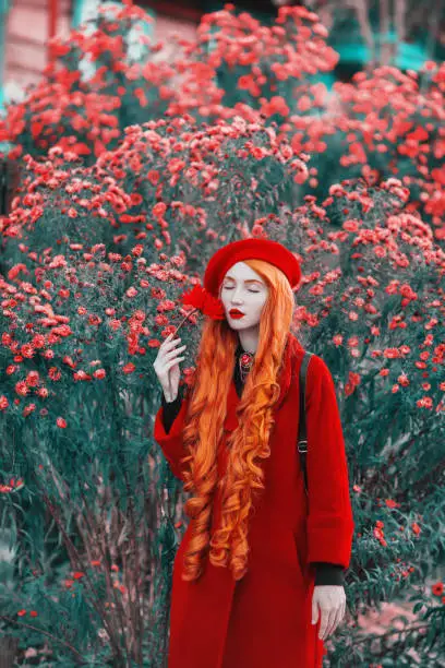 Redhead woman in red coat on garden background. Fashion model with long red hair with flower in hand. Red turban and stylish coat.