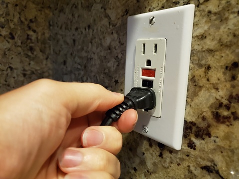 Close-up, personal perspective image of human hand of a man plugging an electrical cord into a Ground Fault Circuit Interrupter (GFCI) electrical outlet on the wall in a domestic room, September 12, 2019