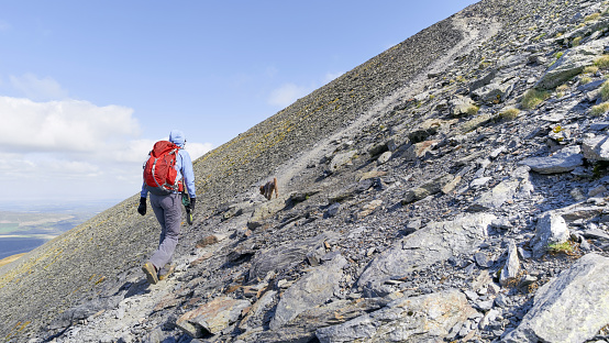 A hiker walking up a steep rocky mountain trail leading to the summit of Skiddaw in the English Lake District.