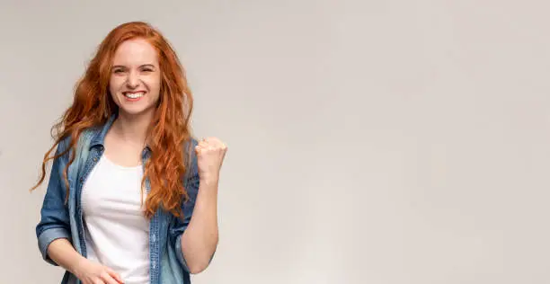 Emotional ginger girl smiling and raising clenched fist in the air, feeling excited, Panorama with copy space