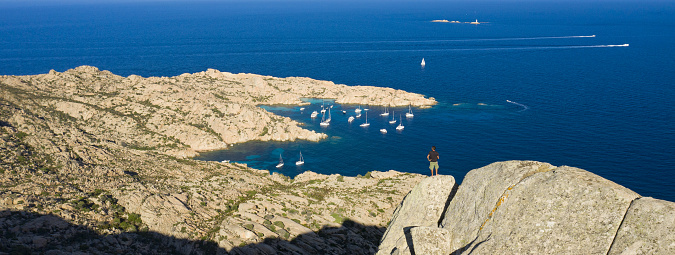 View from above, stunning aerial view of a person on the top of a granite mountain. Cala Coticcio also known as Tahiti in the background. La Maddalena Archipelago, Sardinia, Italy.