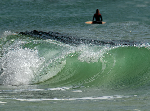 Unrecognizable person in very soft focus sitting on their surf board in the distance with their back to the camera behind a large curling breaking wave that is the main focus in the foreground.