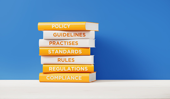 Books of compliance, regulations, rules and guidelines are sitting on top of each other. The books have unique texts on their spines related to compliance subject.