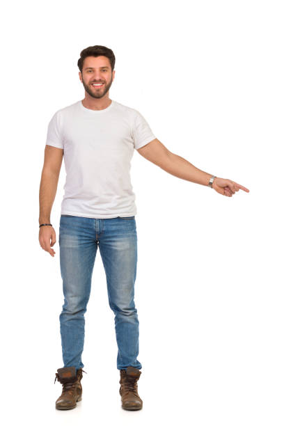 istock Smiling Handsome Man In Jeans And White T-shirt Is Standing, Pointing Down And Looking At Camera. Front View. 1174534636