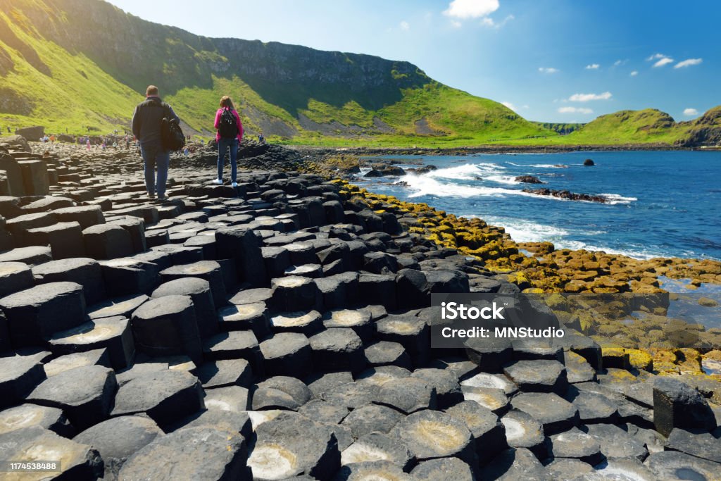 Two tourists walking at Giants Causeway, an area of hexagonal basalt stones, County Antrim, Northern Ireland. Famous tourist attraction, UNESCO World Heritage Site. Two tourists walking at Giants Causeway, an area of hexagonal basalt stones, created by ancient volcanic fissure eruption, County Antrim, Northern Ireland. Famous tourist attraction, UNESCO World Heritage Site. Ireland Stock Photo
