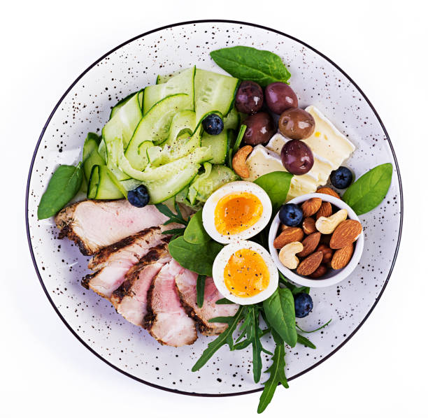 케톤 생성 식단. 케토 브런치. 삶은 달걀, 돼지 고기 스테이크와 올리브, 오이, 시금치, 브리 치즈, 견과류와 블루 베리. 맨 위 보기 - close up green plate salad 뉴스 사진 이미지