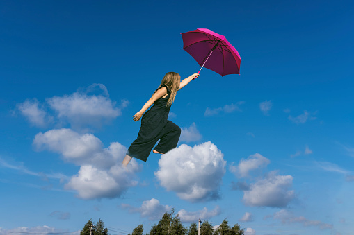 Cute girl jumping funny in clouds above earth. Small cheerful girl walking through sky with pink umbrella. Levitation concept