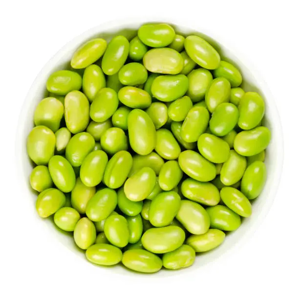 Edamame. Green soybeans in white bowl. Mukimame, unripe soya beans outside the pod. Glycine max, a legume, edible after cooking and a protein source. Closeup, on white background, macro food photo.