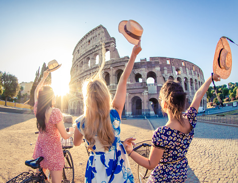 Tres jóvenes felices amigos turistas con bicicletas agitando sombreros en el Coliseo de Roma, Italia al amanecer. photo