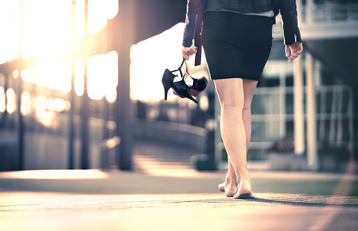 Woman holding high heels in hand and walking home from party barefoot. Businesswoman took off uncomfortable shoes. Lady with stilettos and miniskirt at sunrise or sunset in city street.