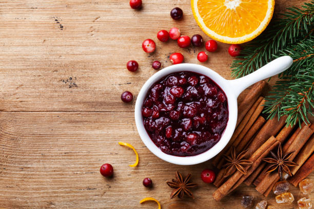 Cranberry sauce in ceramic saucepan with ingredients for cooking decorated with fir tree for Christmas or Thanksgiving day on rustic kitchen table. Top view. stock photo