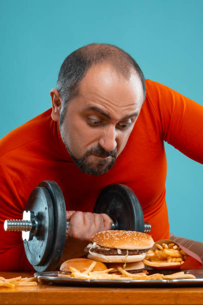 istock Close-up portrait of a middle-aged man with beard, dressed in a red turtleneck, posing with burgers and french fries. Blue background. Fast food 1174647513